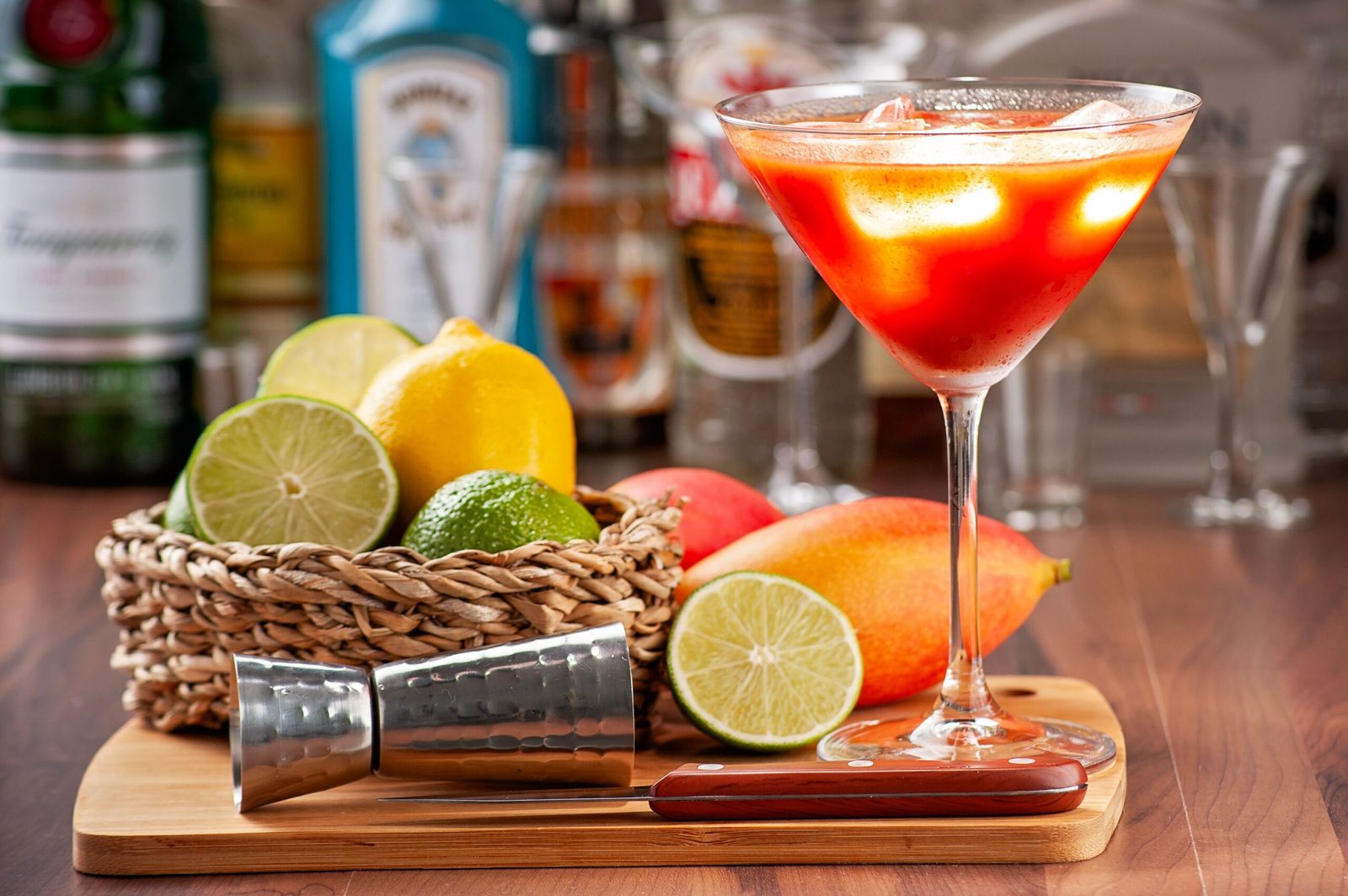 verre à cocktail rempli d'un cocktail coloré avec des fruits sur les coté dans une corbeille et des bouteille de spiritueux en fond d'image